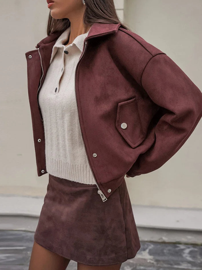 Woman wearing a burgundy jacket over a white sweater and skirt, standing against a neutral background.